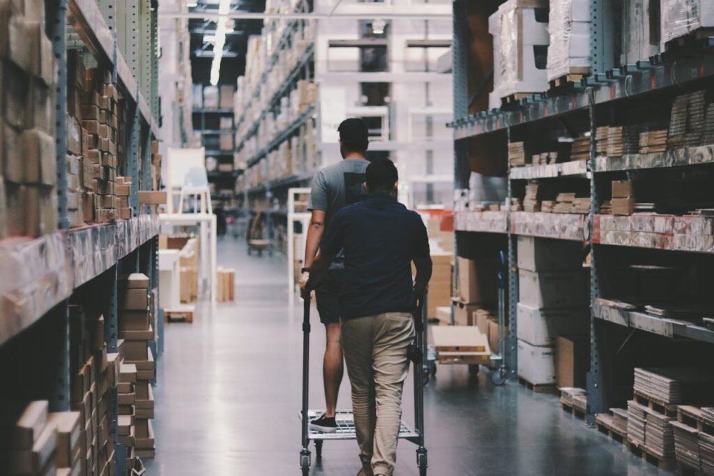 pexels photo 1797428 Two men maneuver a trolley in a large warehouse filled with boxes and shelves.